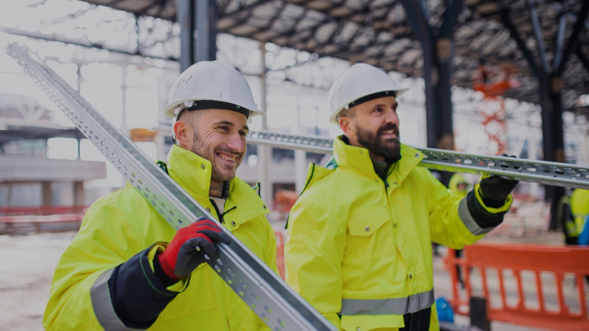 Deux ouvriers souriant sur un chantier en extérieur, portant des équipements de protection, dont l’un transporte du matériel sur son épaule.