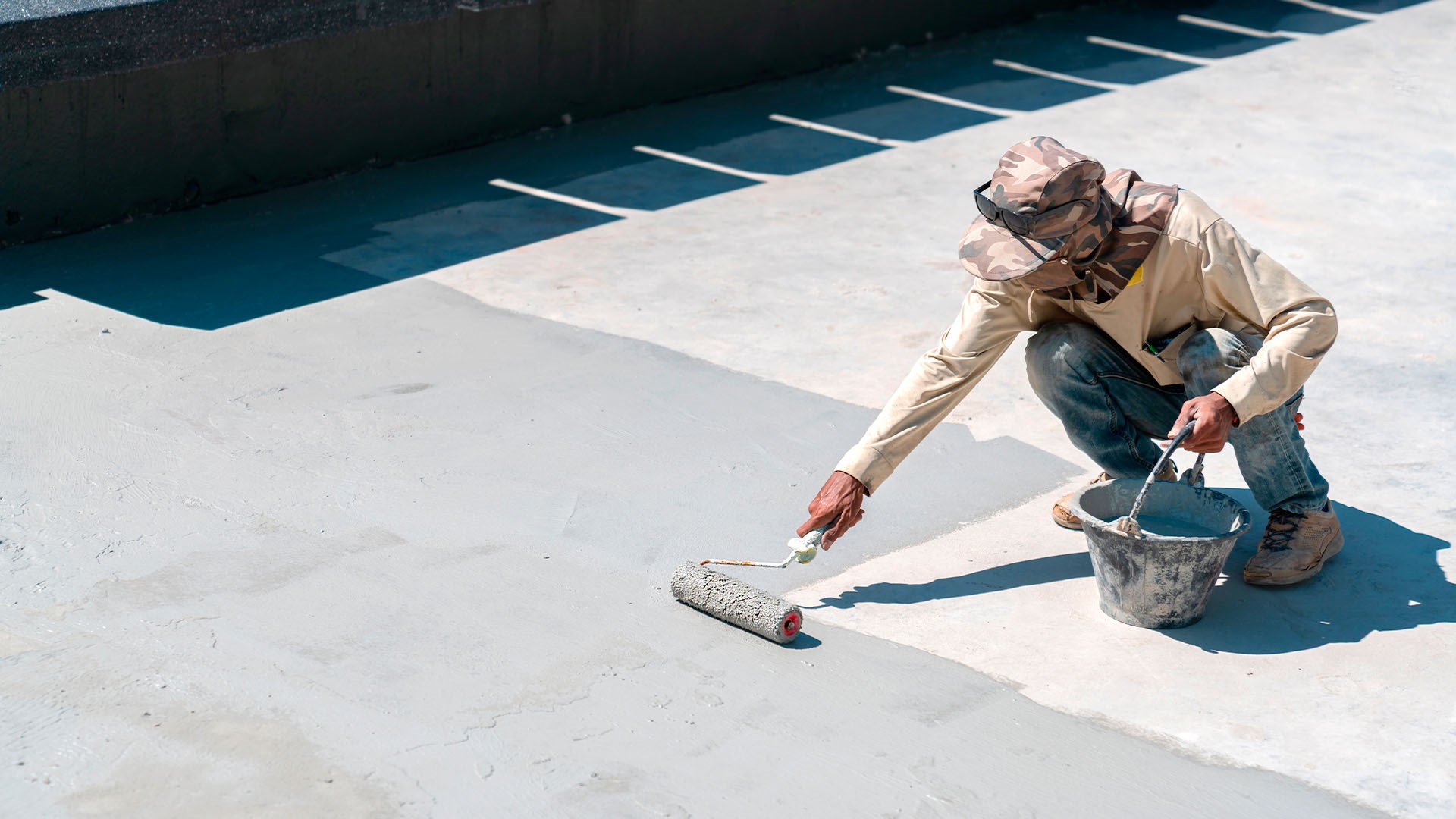 Travailleur appliquant une couche de béton sur une surface extérieure, à l’aide d’un rouleau fixé à un manche long, avec un seau à portée de main.